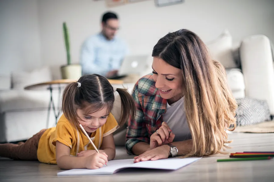 A mother and her young daughter laying on the floor together as the daughter learns to write, holding a pen and drawing in a notebook. In the background, the father works on his laptop.
