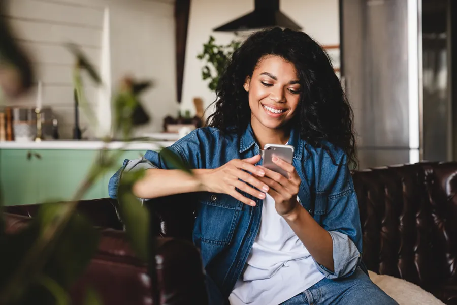 A young woman sitting on the leather couch in her living room, holding her smartphone and smiling.