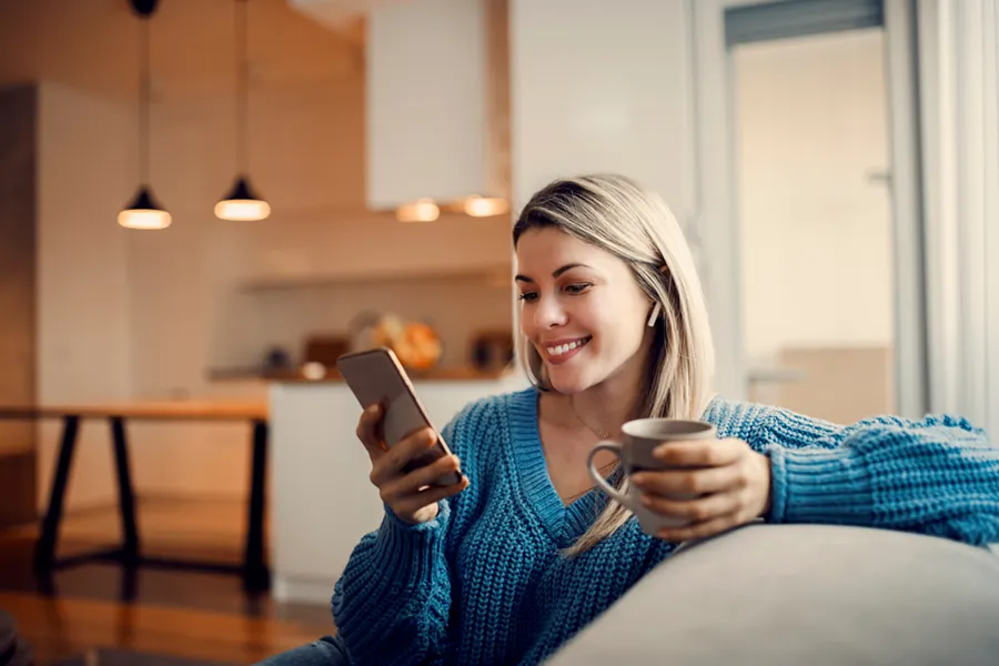 Young woman lounging on her couch with a hot beverage, looking at something on her smartphone and smiling.