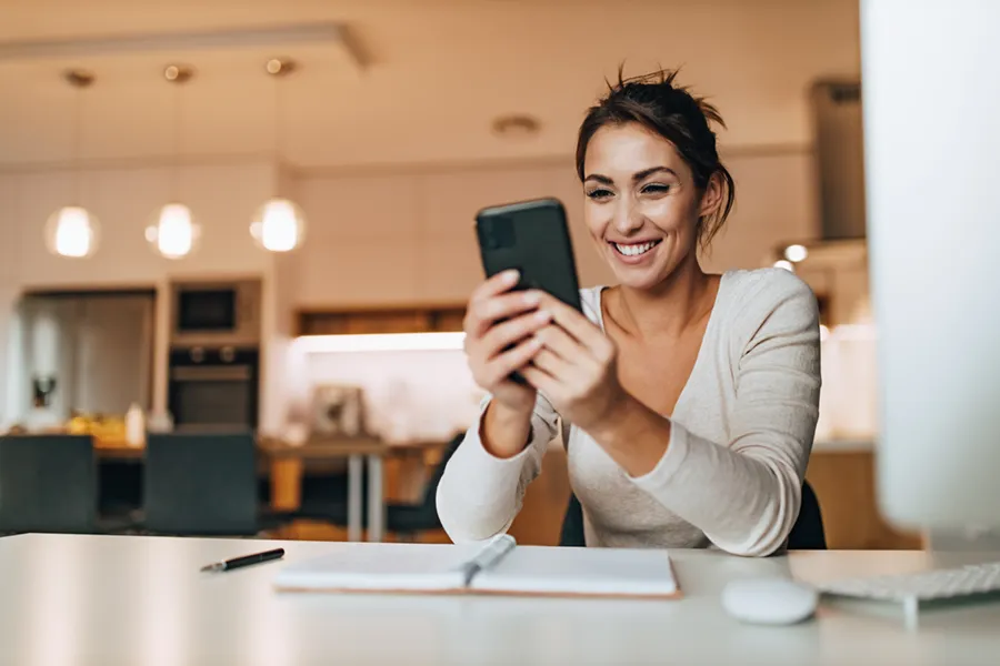 A young woman using her smartphone and sitting at her dining table near her kitchen with a notebook and pen open before her.